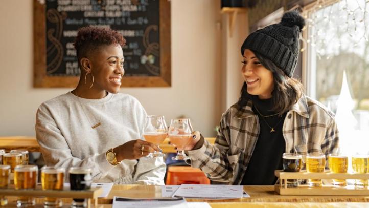 Two women cheers with goblets. They are sitting at a table at Boxing Rock Brewing Co. and each has a flight of beer in front of them.