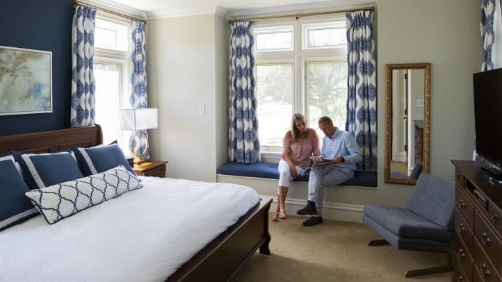 A man and a woman sit on a window bench in a hotel room. There is a bed against the wall facing a dresser with a TV on top.